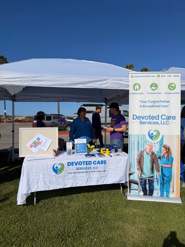 Outdoor information booth under a white canopy with Devoted Care banners and staff.