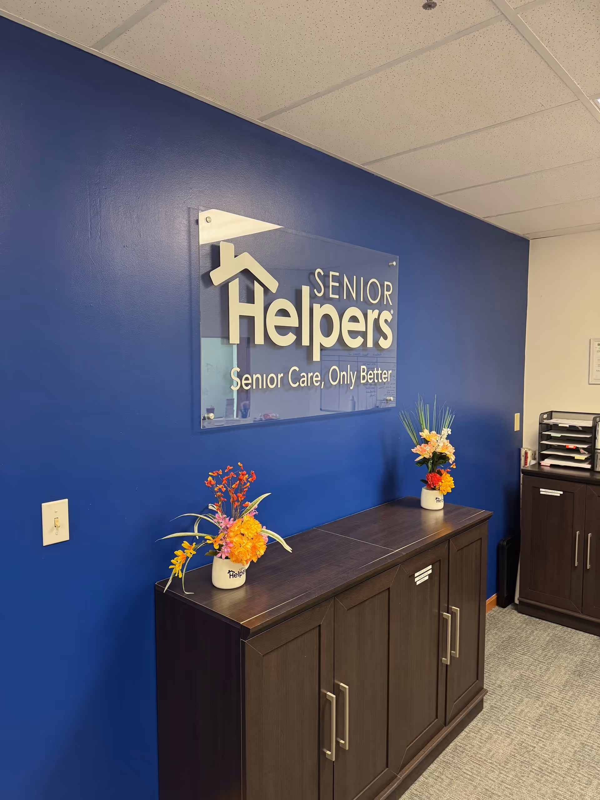 Blue interior wall with Senior Helpers sign, dark cabinets, and flower arrangements on top.