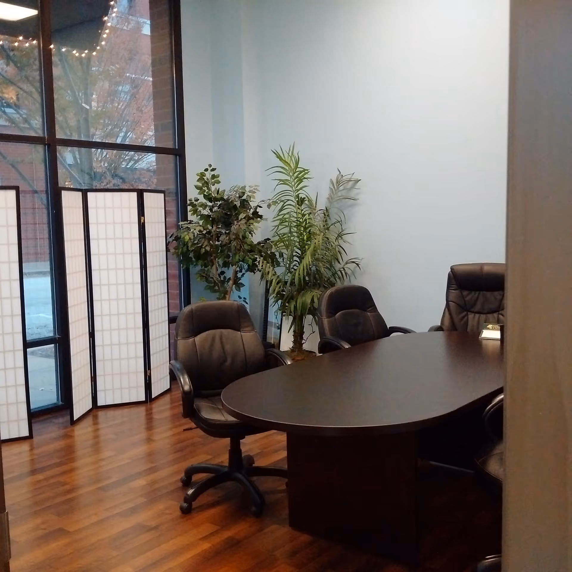 Office conference room with dark wood table, black chairs, plants, and divider screens.