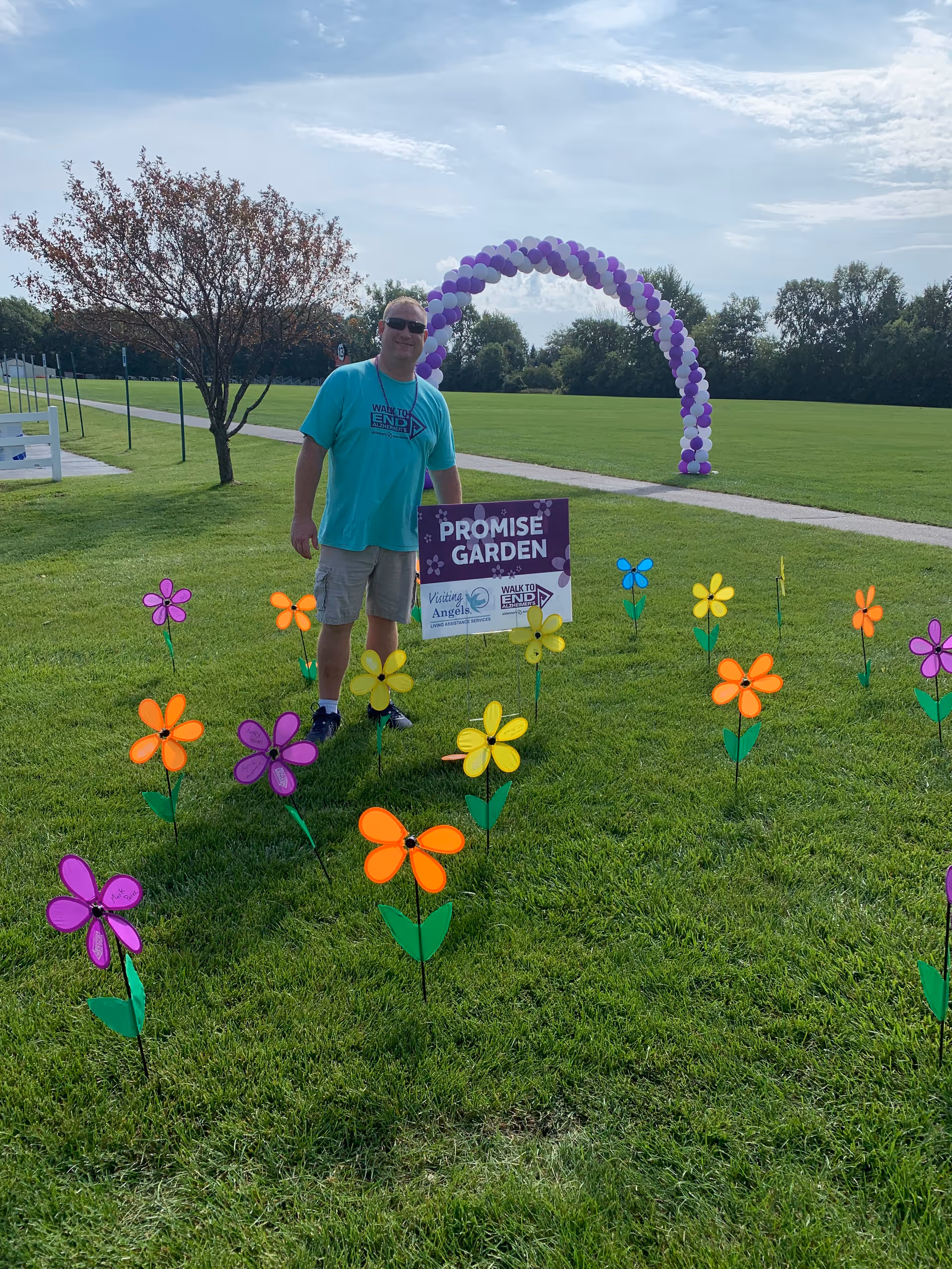 Man stands in a grassy park with a purple balloon arch, colorful flowers, and a sign.