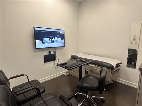 Small medical examination room featuring an examination table, desk, monitor, and several chairs.