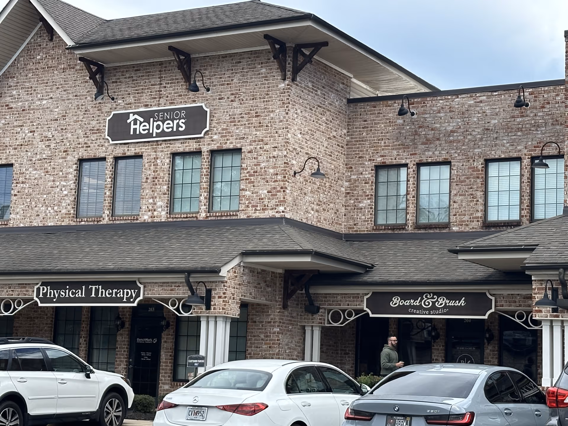 Brick building facade with Senior Helpers sign, storefronts Physical Therapy and Board & Brush, parked cars.