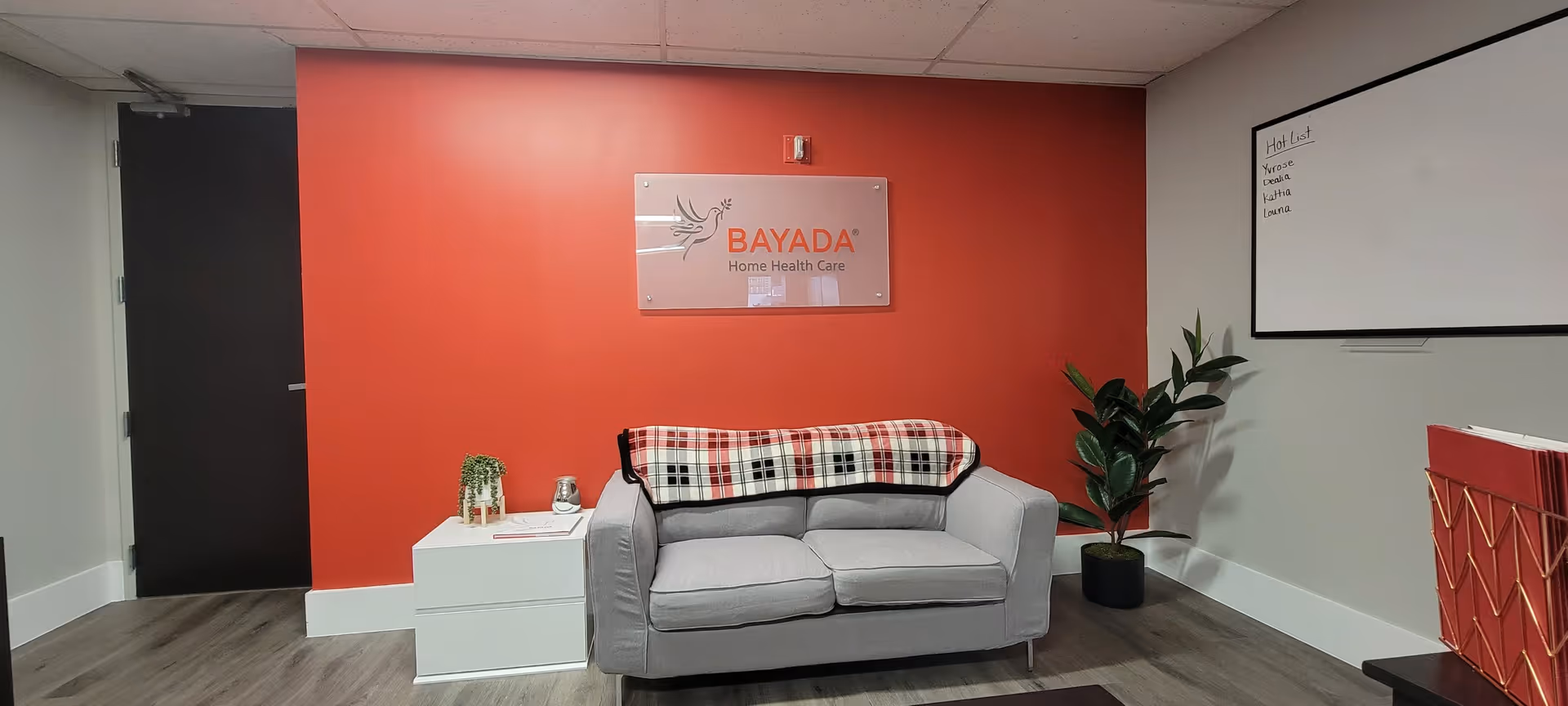 Reception-style lounge with a gray sofa, red accent wall, BAYADA sign, and plants.