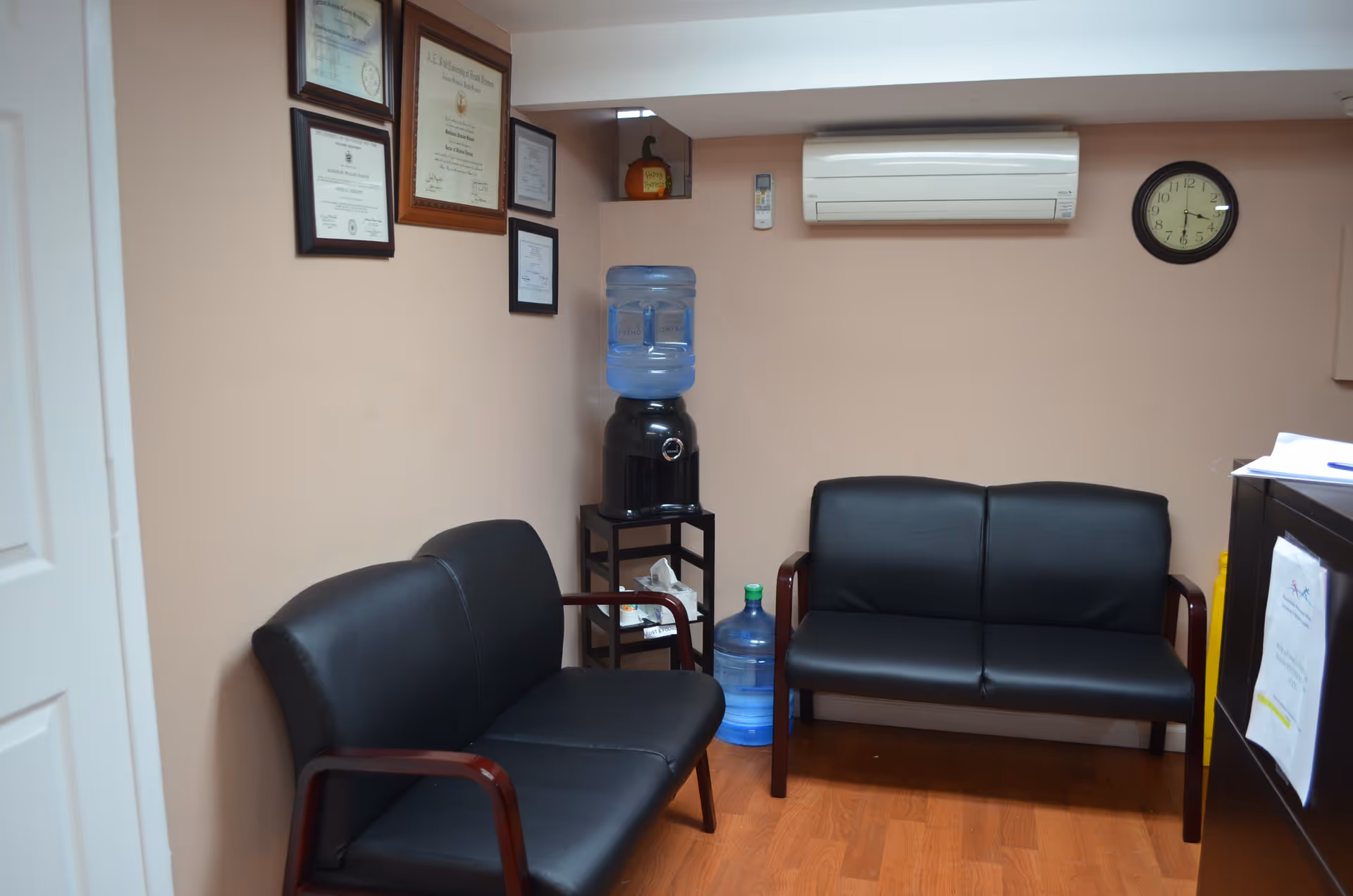 Interior waiting area with black leather chairs, a water cooler, clock, and framed certificates.