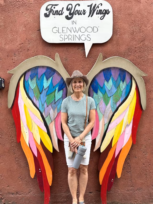 A smiling woman stands before a large colorful wings mural on an exterior wall.