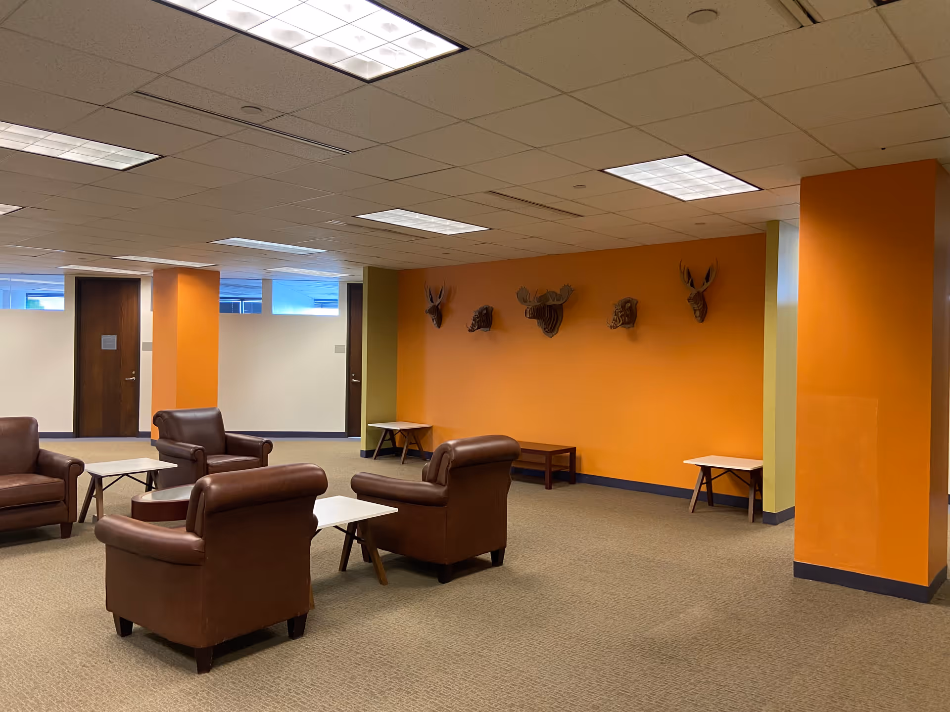 Spacious lounge area with orange walls, brown leather chairs, small tables, and deer head wall art.