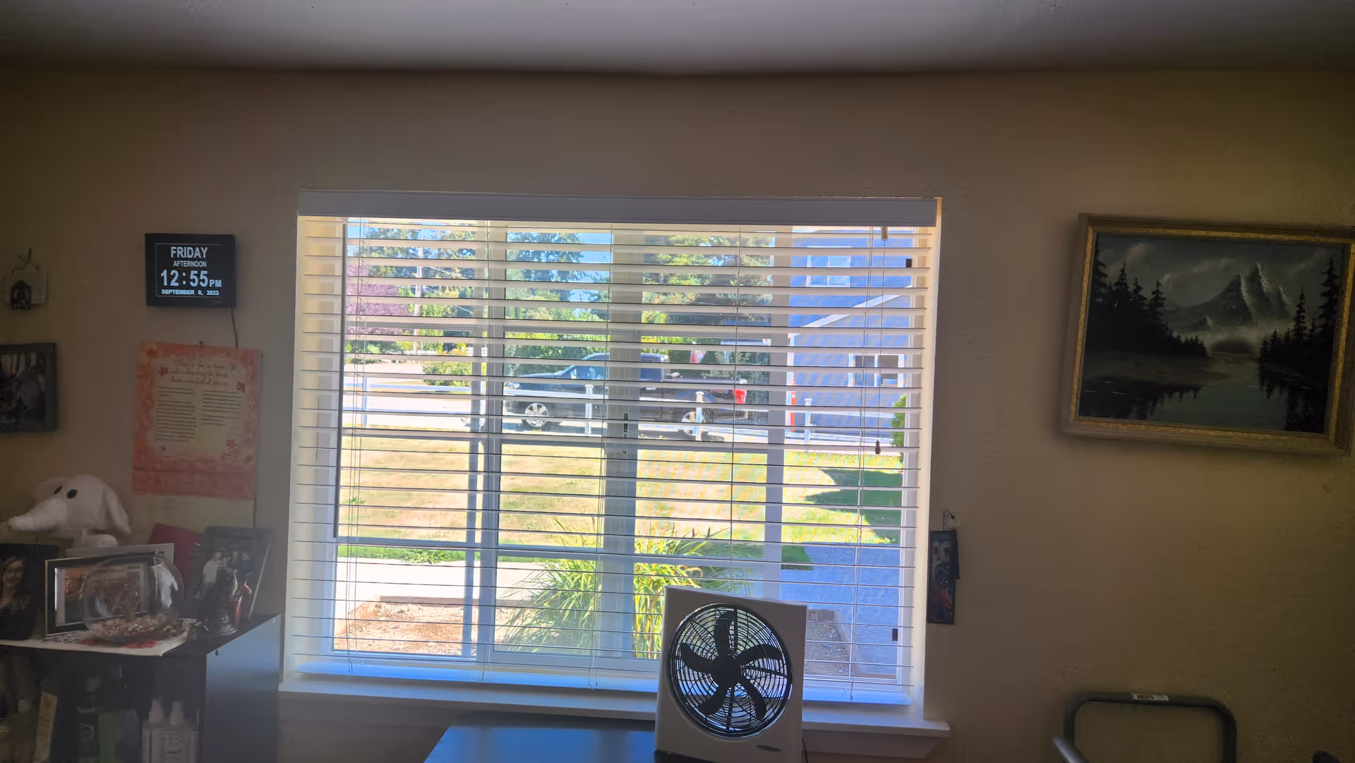 Interior room with large window blinds, a fan on the desk, framed photos, and outdoor view.