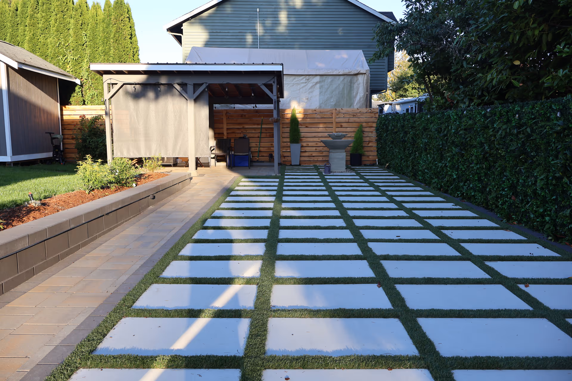 Long outdoor backyard with square stepping stones, a covered patio, fence, and hedge-lined perimeter.