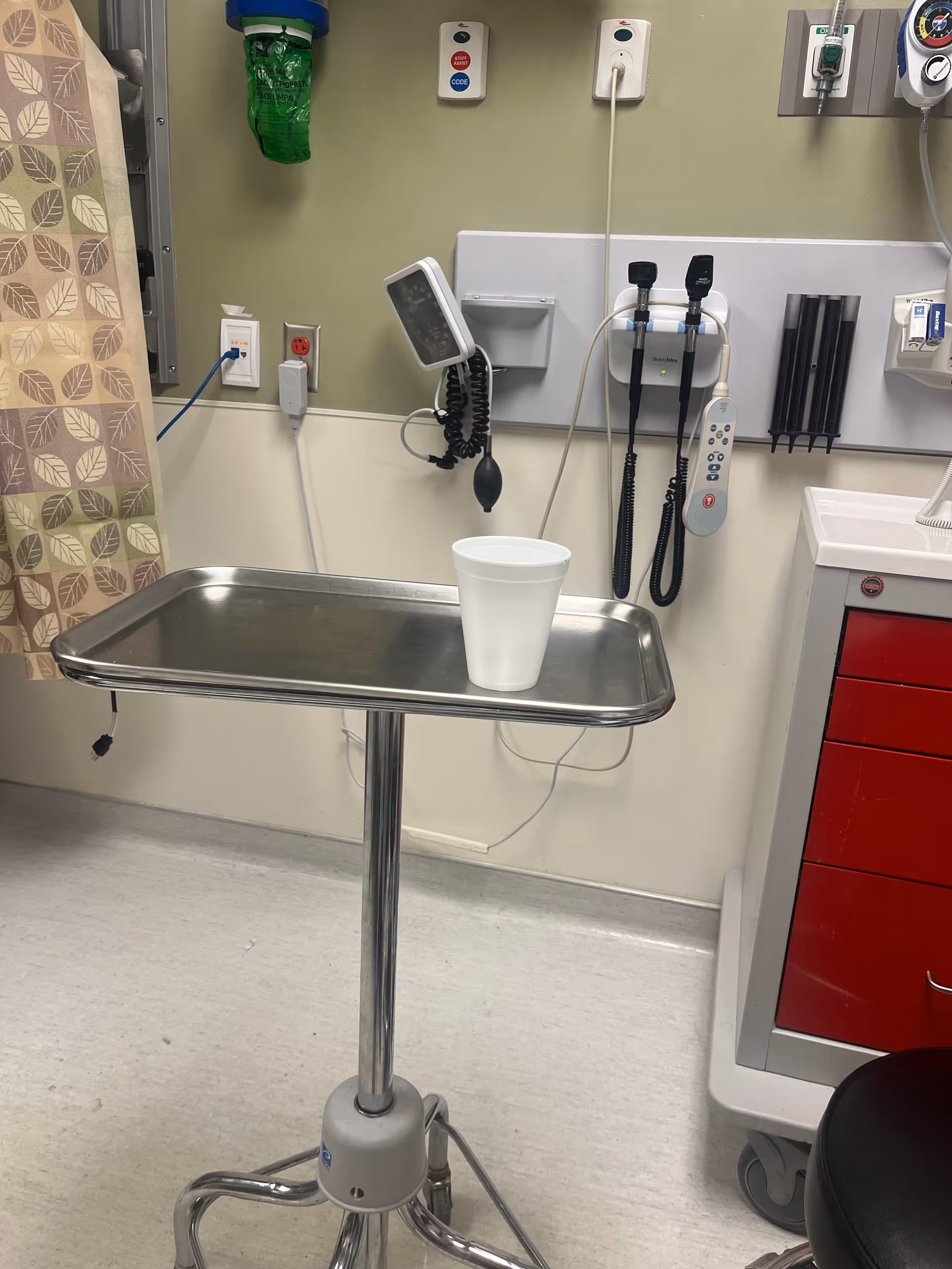 A stainless steel medical tray table with a white cup in a clinical room.