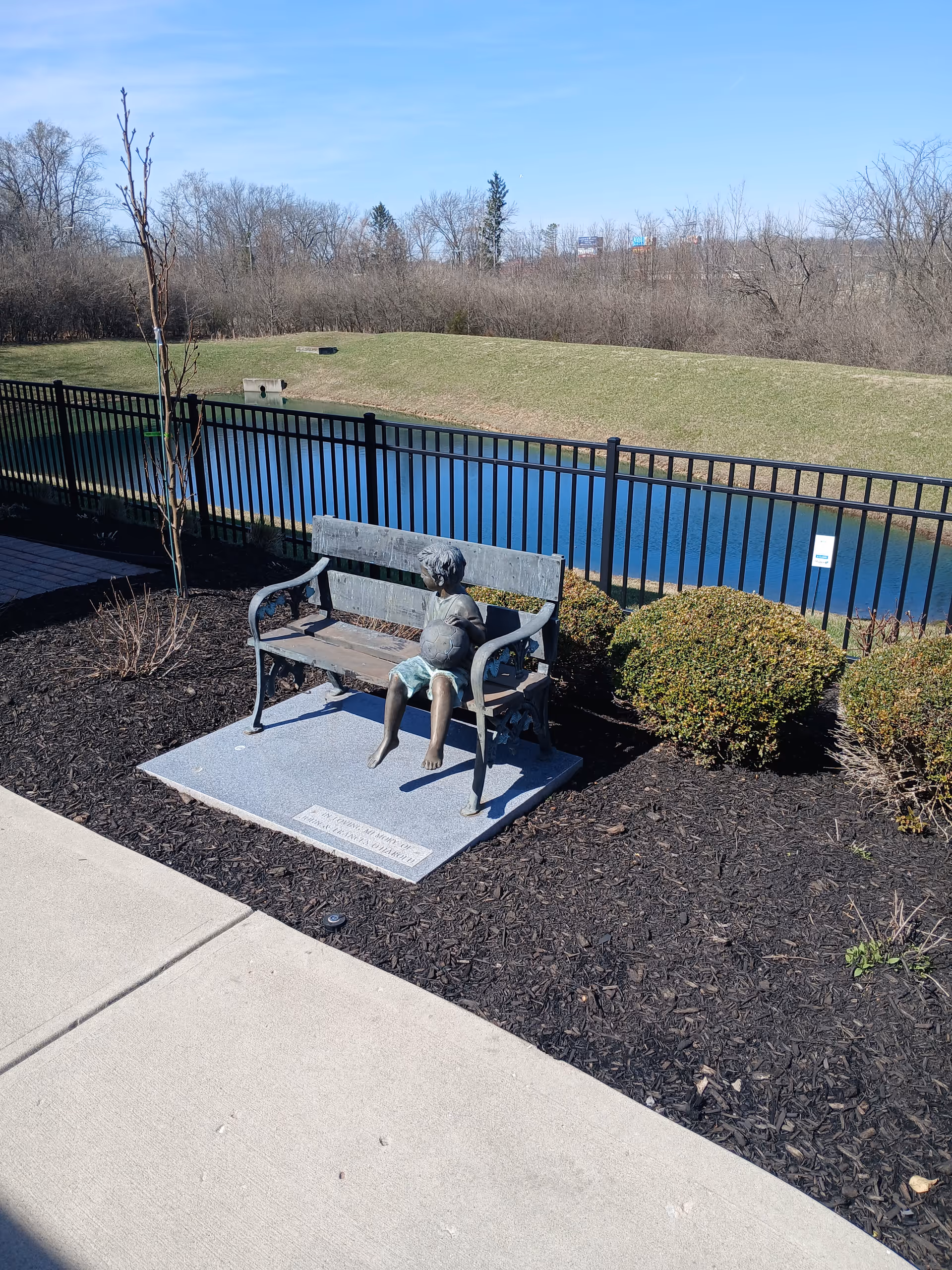 Outdoor bench with statue of a person sits by a black metal fence overlooking a pond.