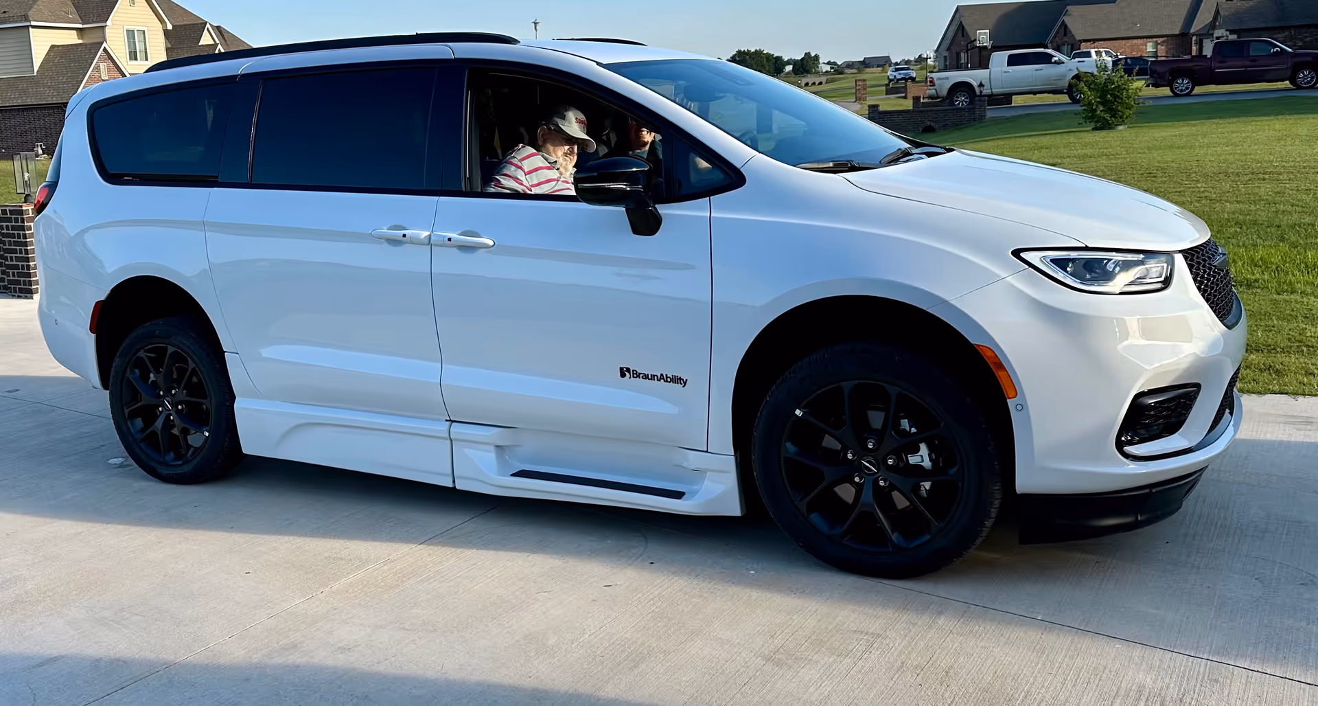 White mobility van parked on driveway with a person in the driver's seat, suburban backdrop.