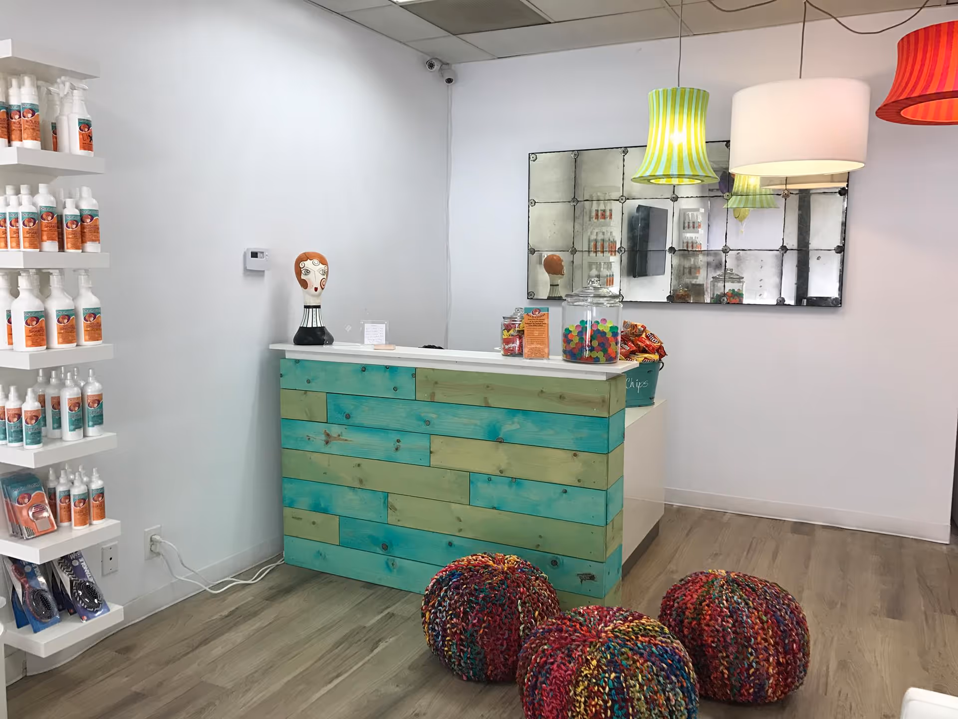 Reception desk with turquoise panels, shelves of bottles, mirrored back wall, and colorful floor poufs.