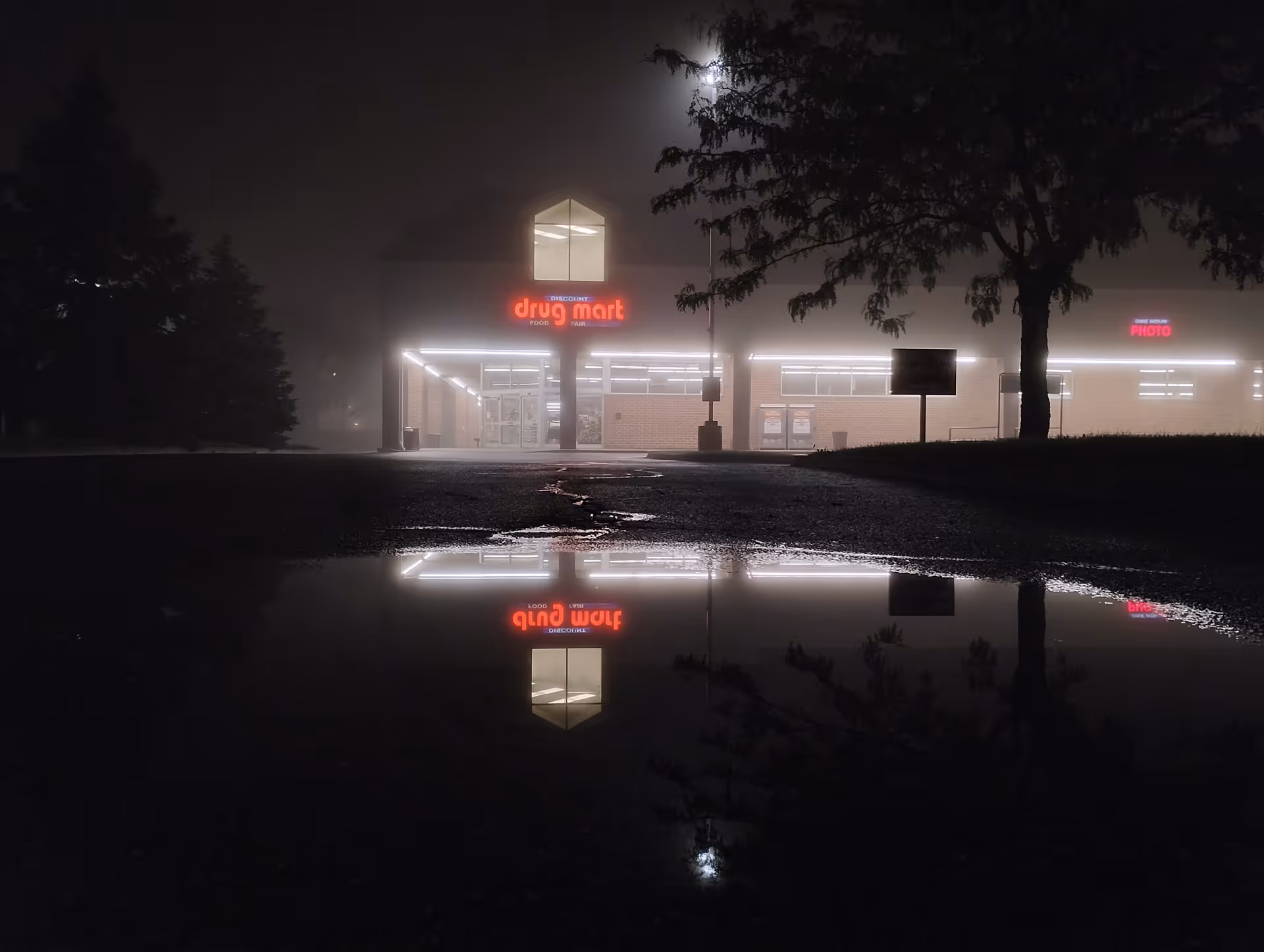 Nighttime exterior of Discount Drug Mart with a glowing sign and its reflection in a puddle.
