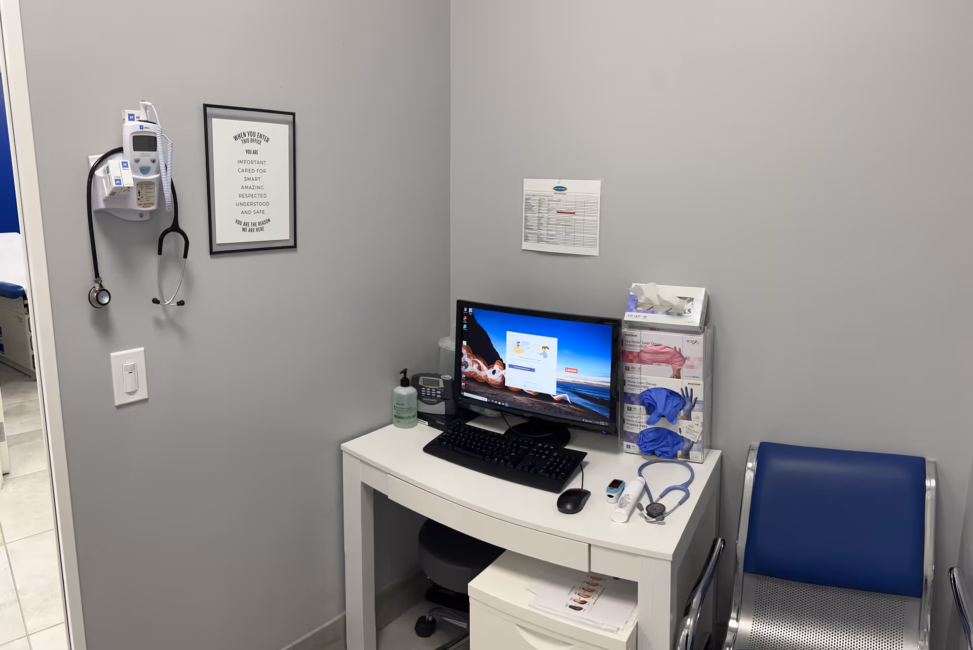 Small medical desk with computer, stethoscope, supplies, and blue chair in a gray-walled room.