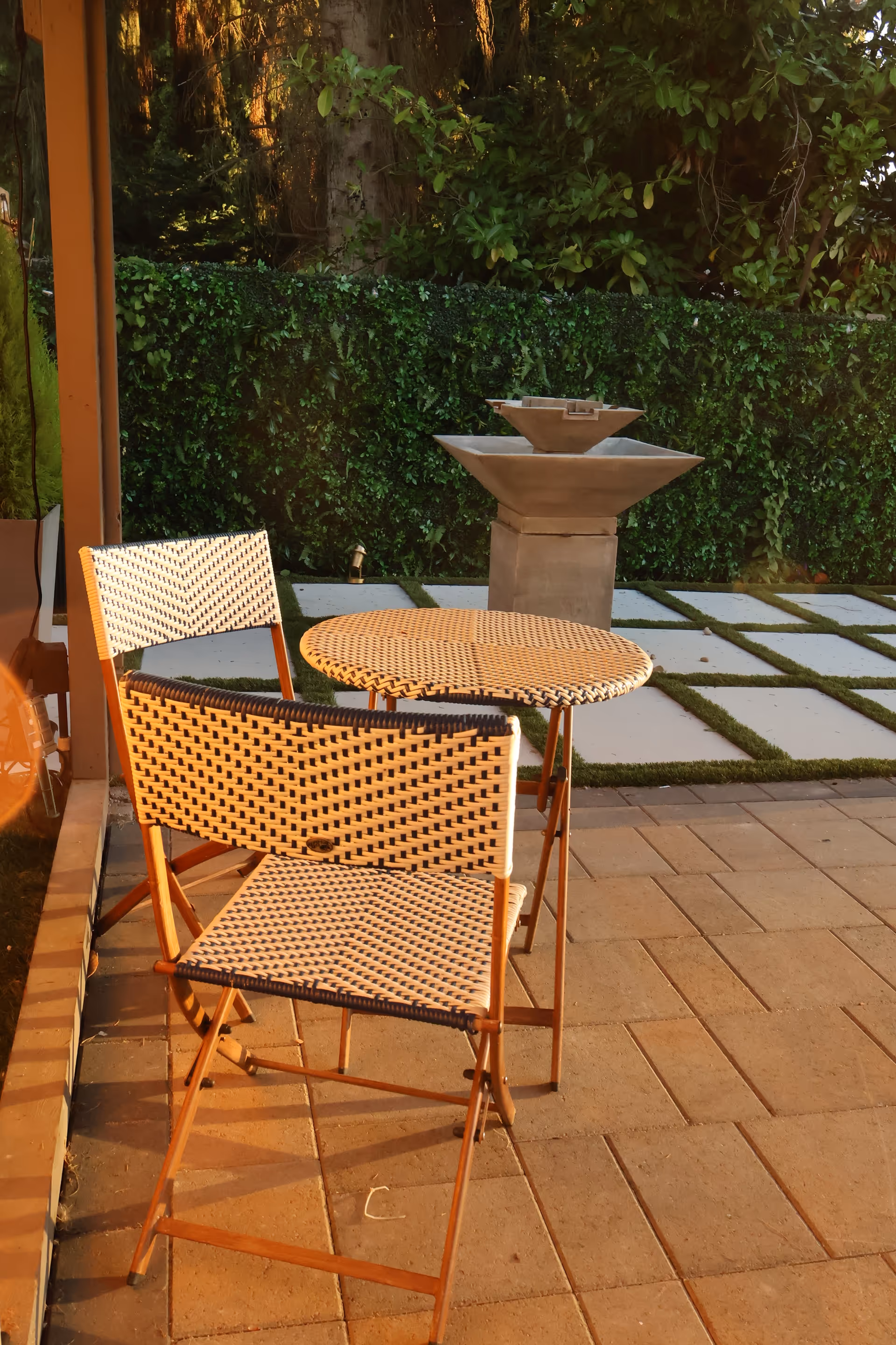 Wicker chairs and a round table on a paved outdoor patio with a fountain and backdrop.