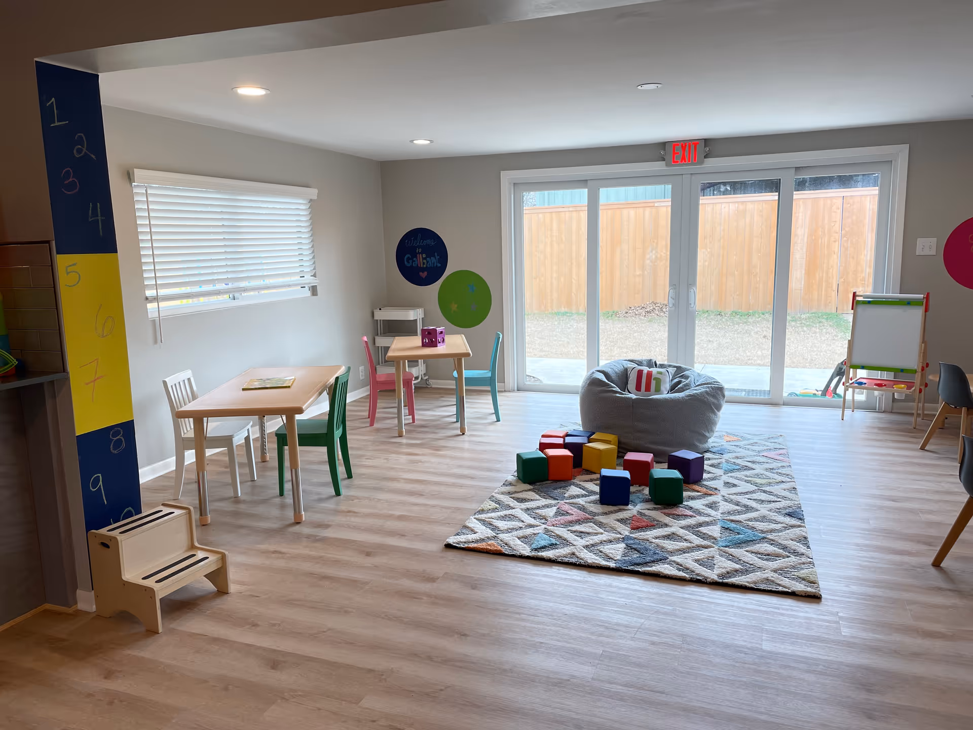 Bright children's playroom with small tables, colorful chairs, a rug, and building blocks.