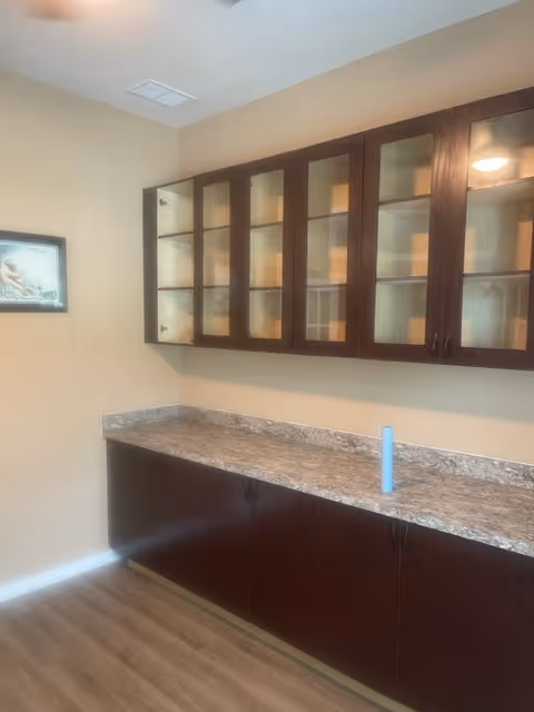 A kitchenette with dark wood cabinets, glass-front uppers, and a granite countertop.