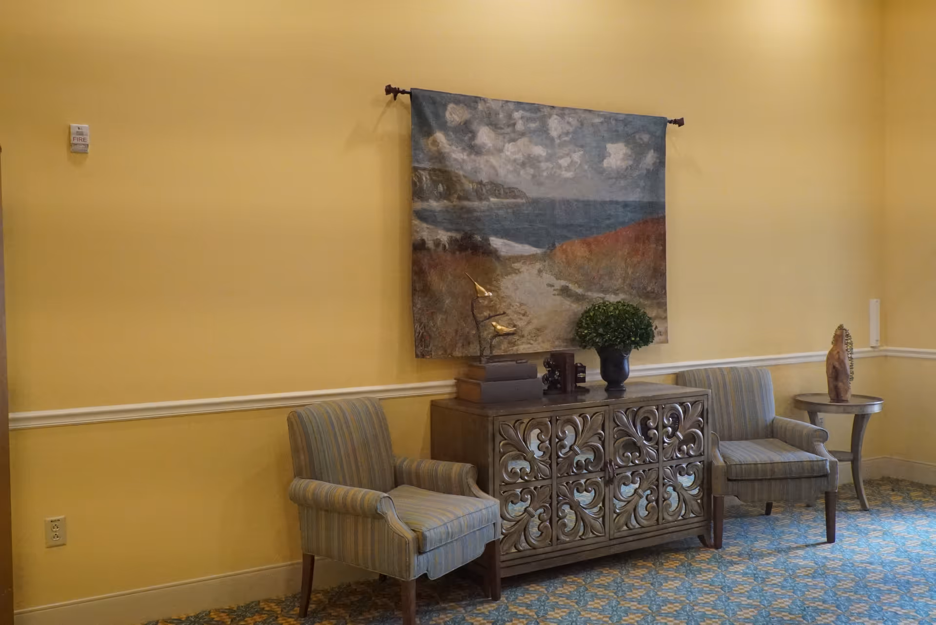 Yellow-walled senior living lobby with two striped chairs, ornate cabinet, and wall art.