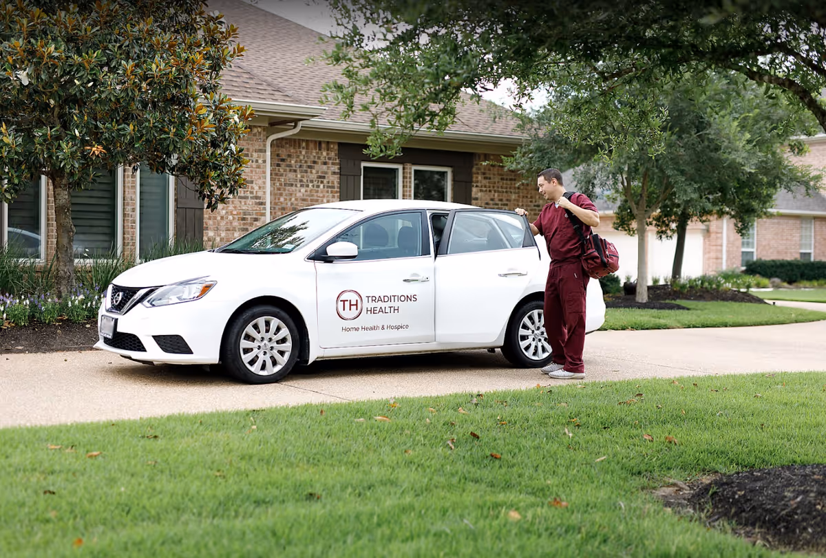 A caregiver in maroon scrubs exits a white Traditions Health car beside a brick facility.