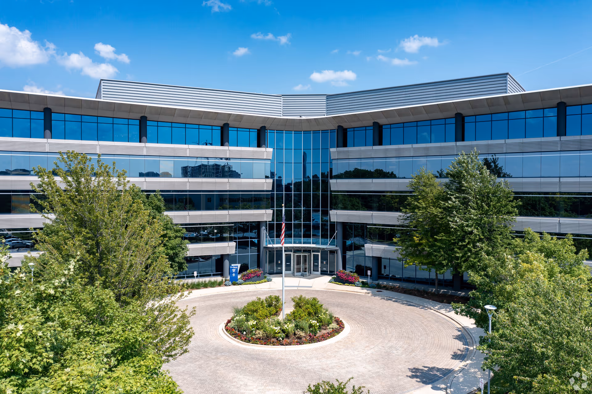 Modern glass-front building with circular driveway and landscaped entrance under blue sky.