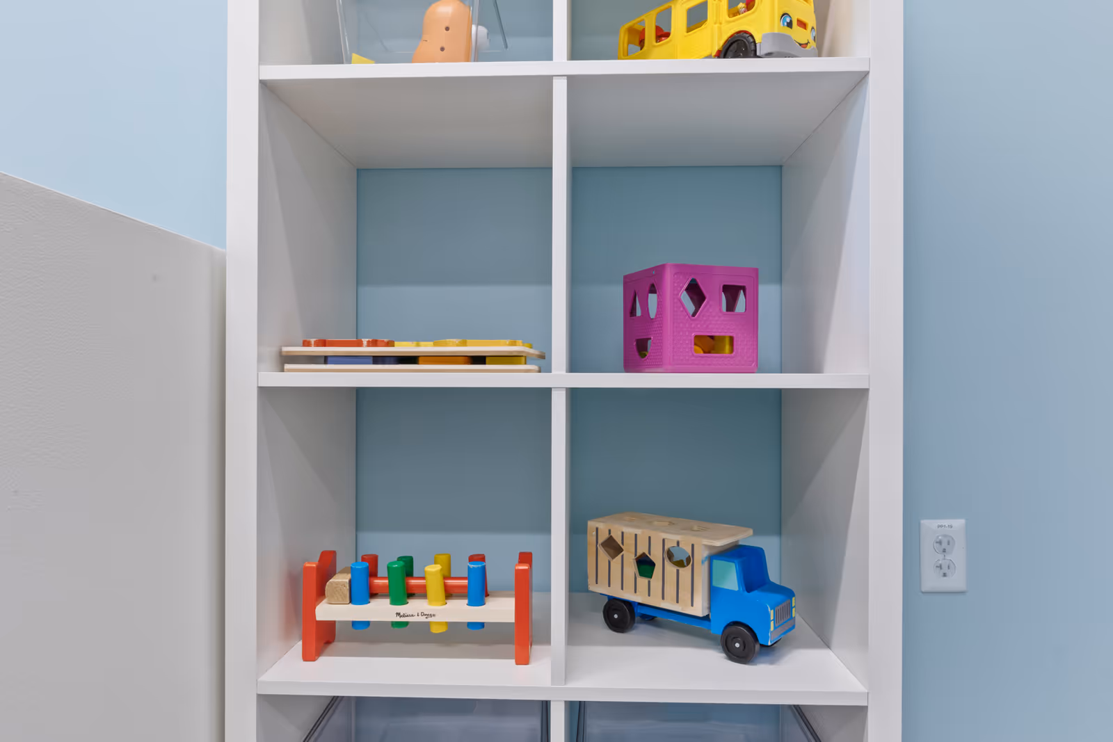 A white shelving unit with colorful wooden toys on blue walls in an indoor play area.