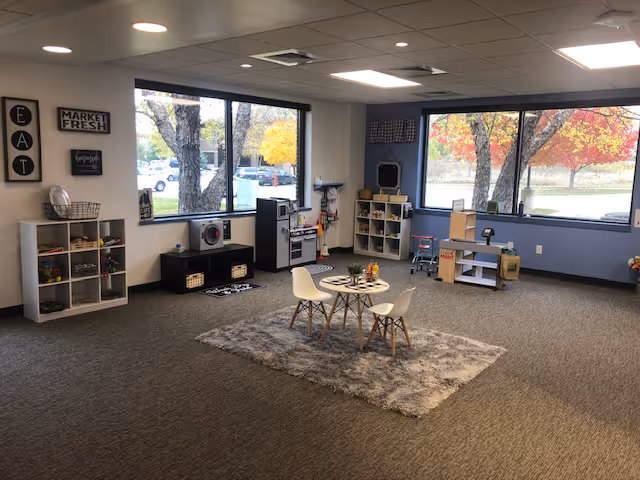 A spacious activity room with toy shelves, a small table and chairs, and large windows.