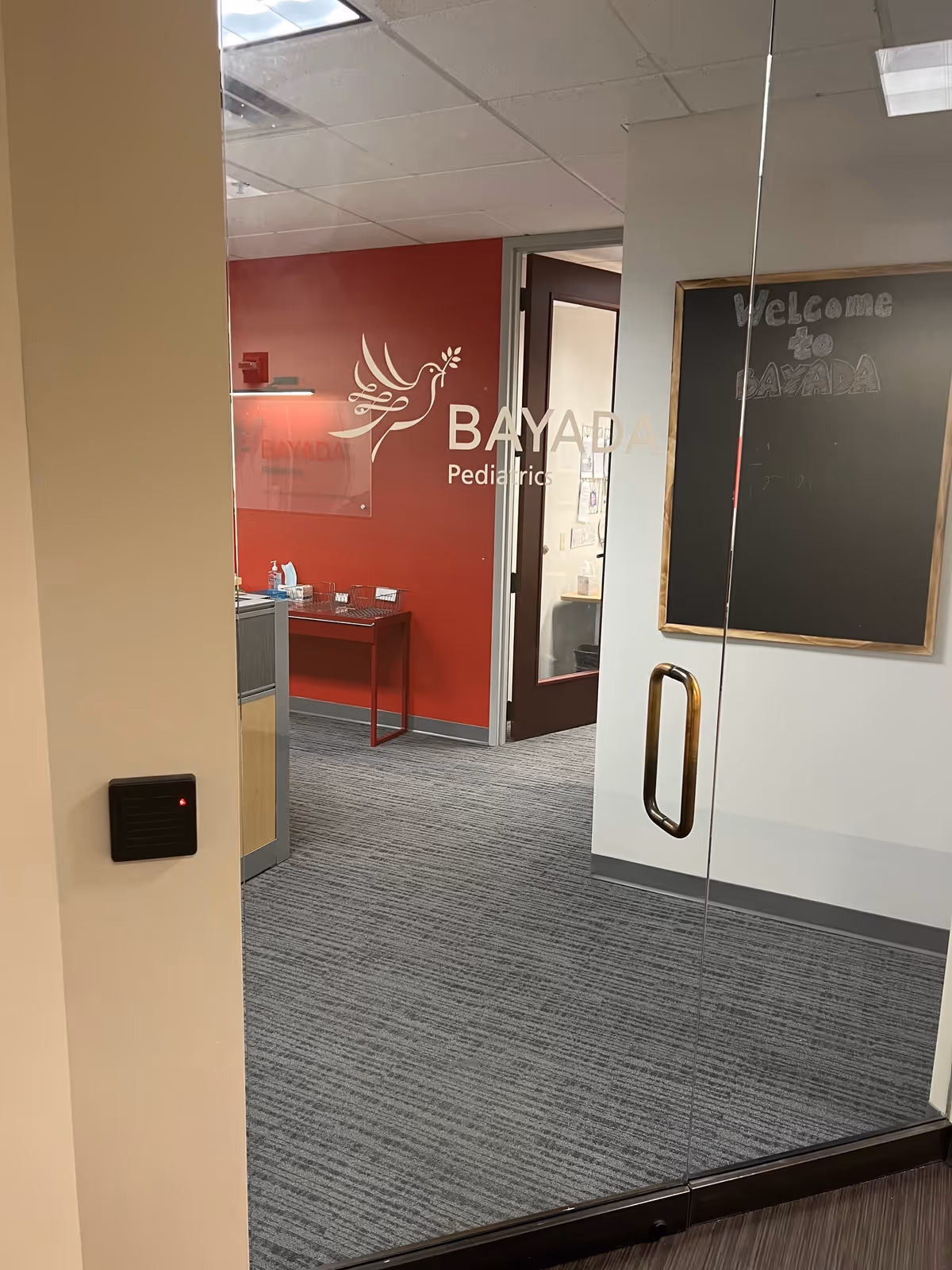 Interior BAYADA Pediatrics reception area with glass entry doors, red wall sign, and a desk.