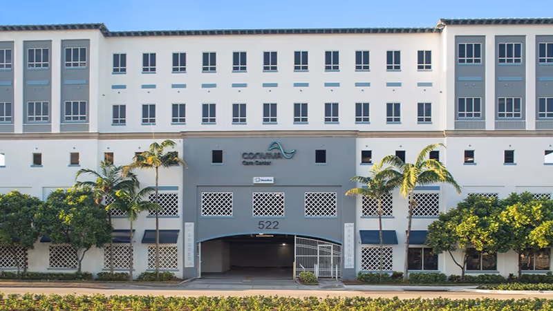 Exterior view of a multi-story senior living facility with palm trees and a central entrance.