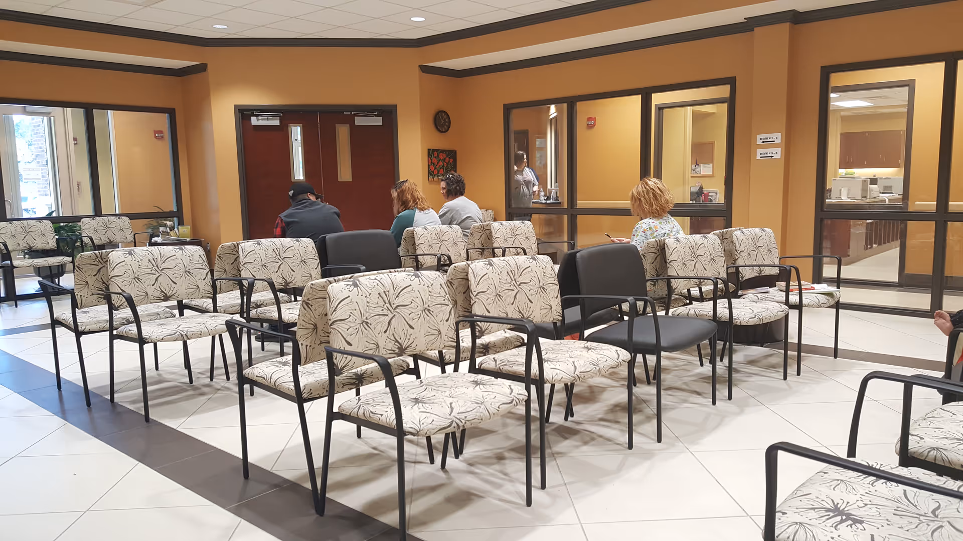 Indoor waiting area with patterned chairs and several people seated near windows.