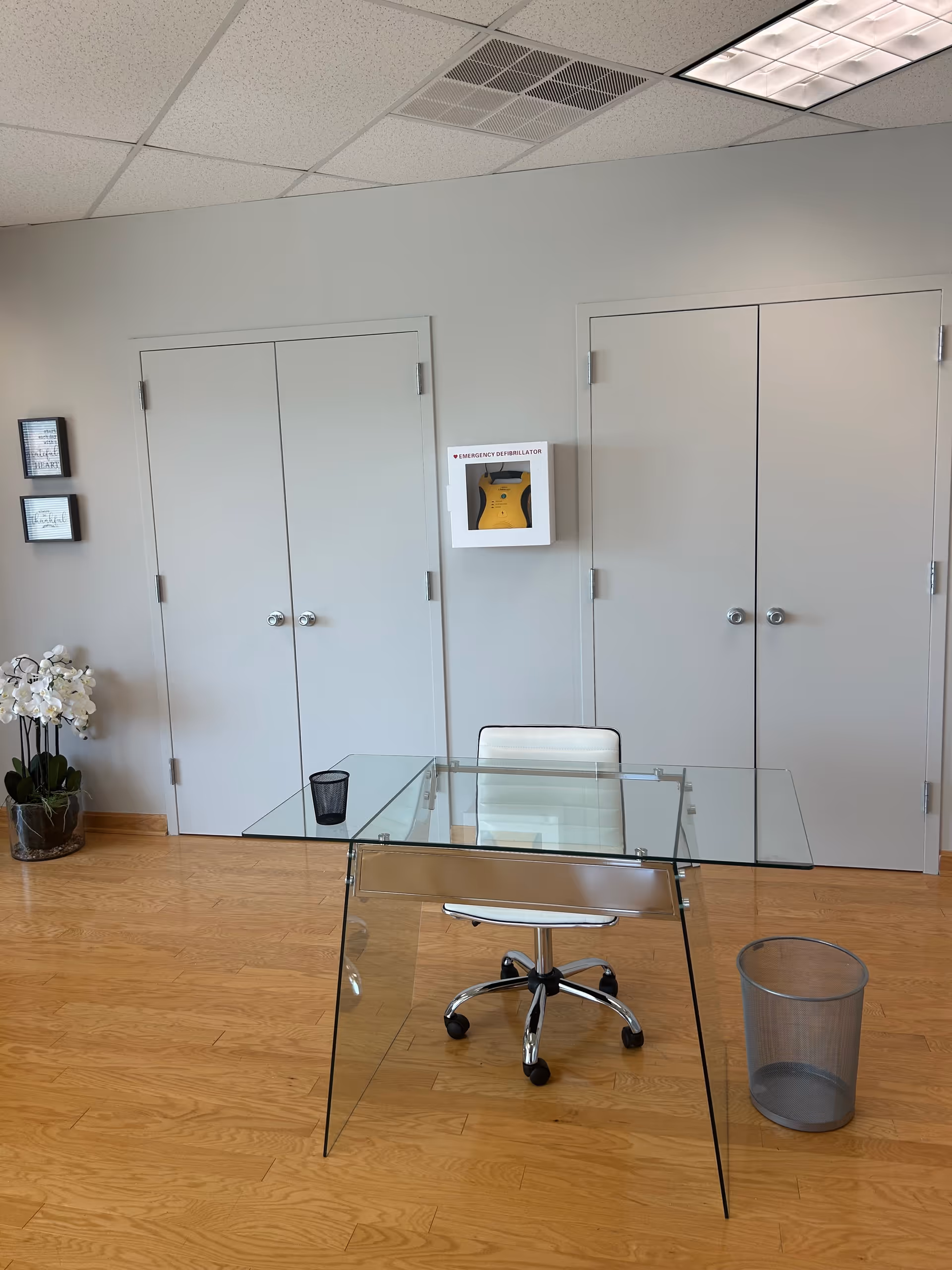 Glass desk with white chair in a modern interior, doors in the background, trash bin nearby.