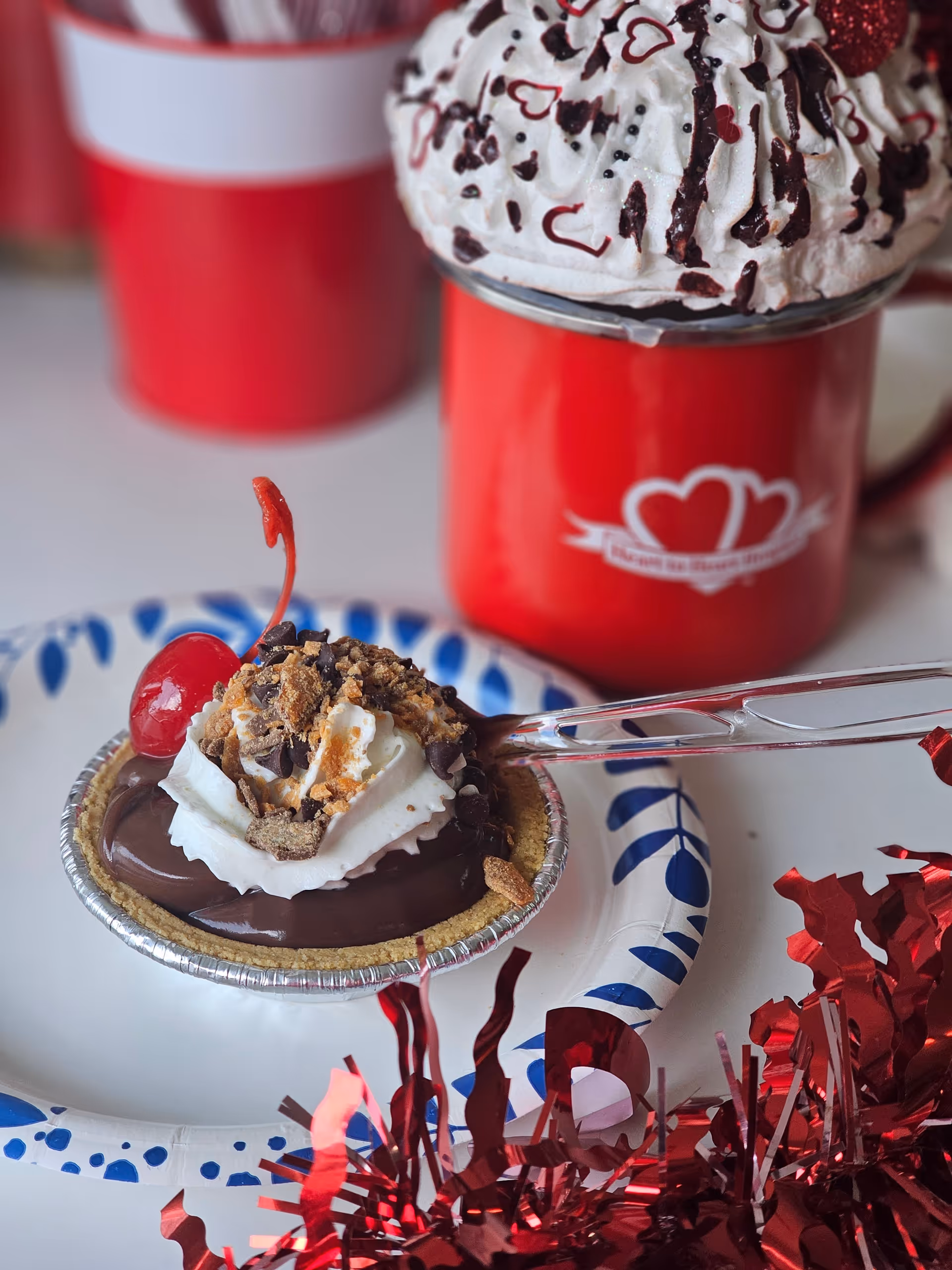 Close-up of a dessert tart topped with whipped cream and a cherry beside red heart mugs.