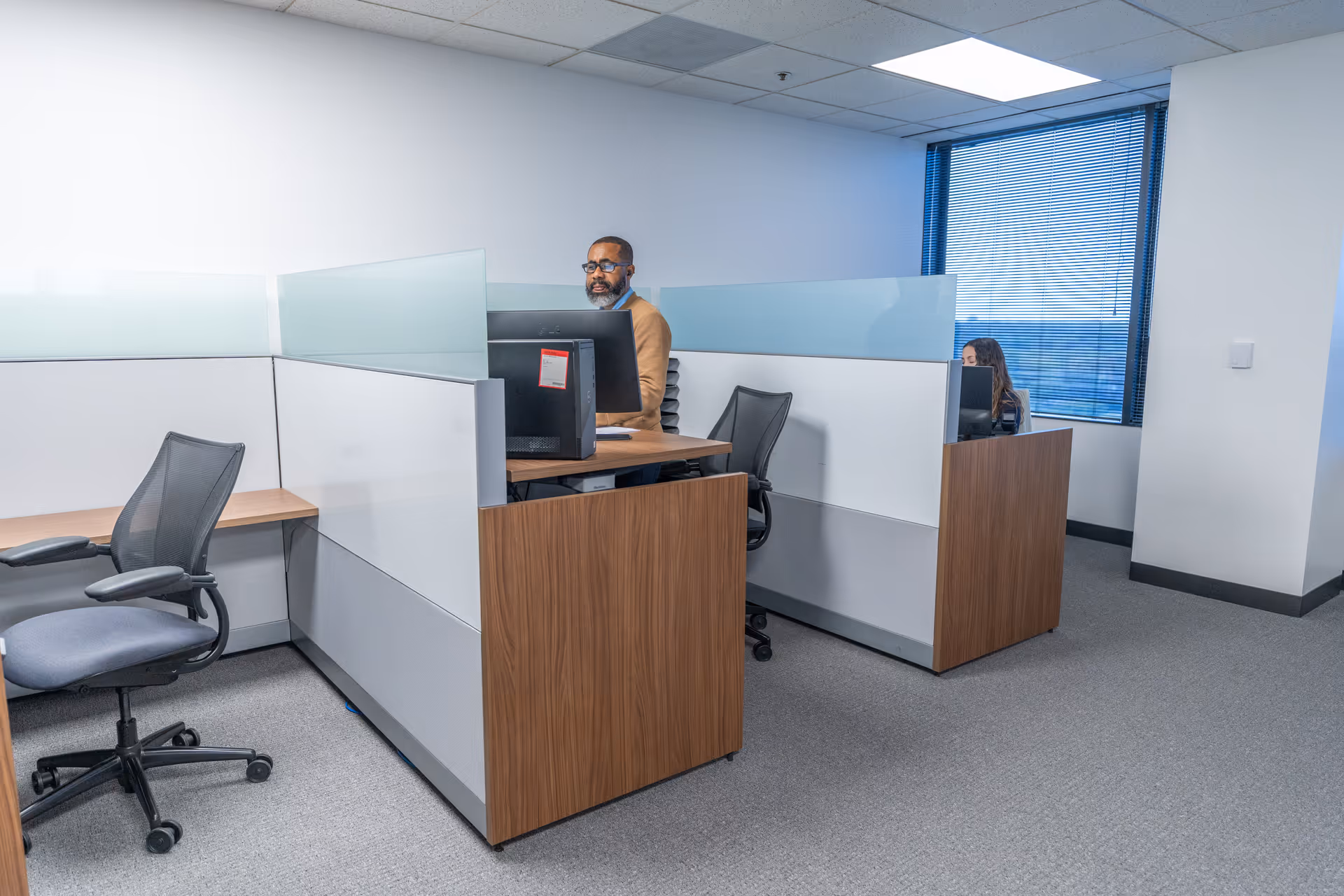 Two office workers sit at cubicle desks in a bright, modern workspace.