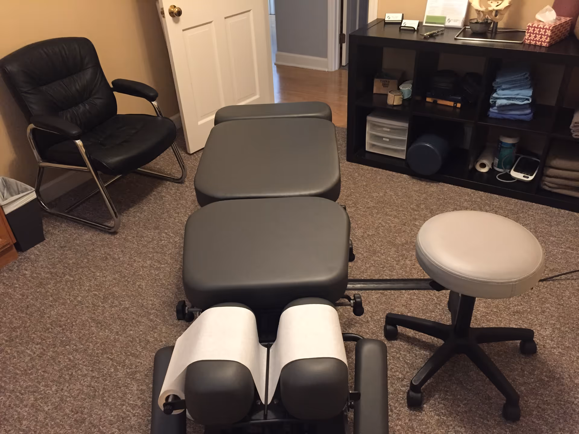 Treatment table with leg supports and a rolling stool in a clinic room.