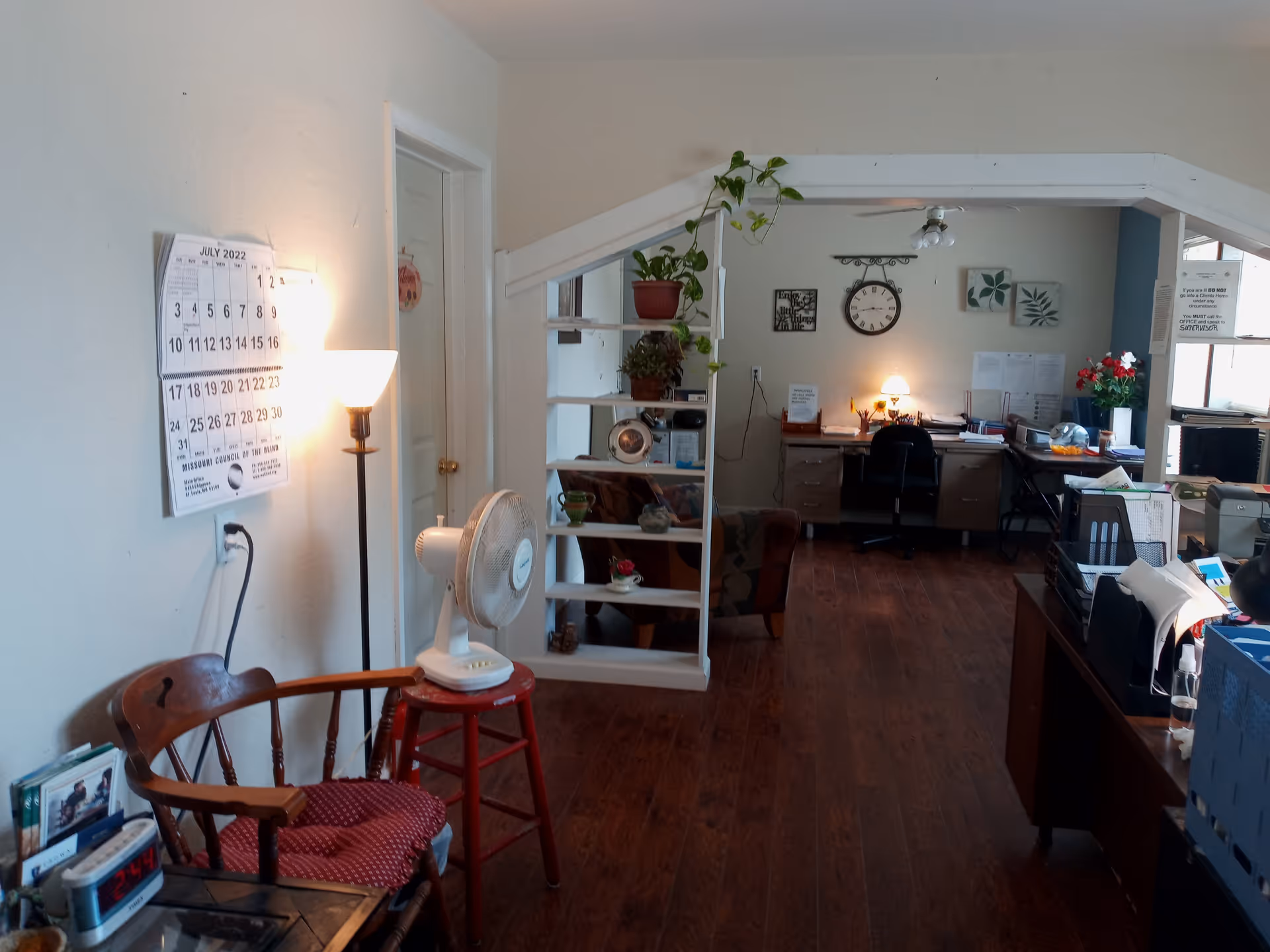 Office area inside a senior facility with a partition shelf, desk, calendar, and standing fan.
