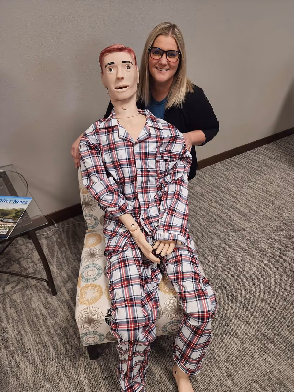 Caregiver stands behind a life-size mannequin in plaid pajamas indoors.