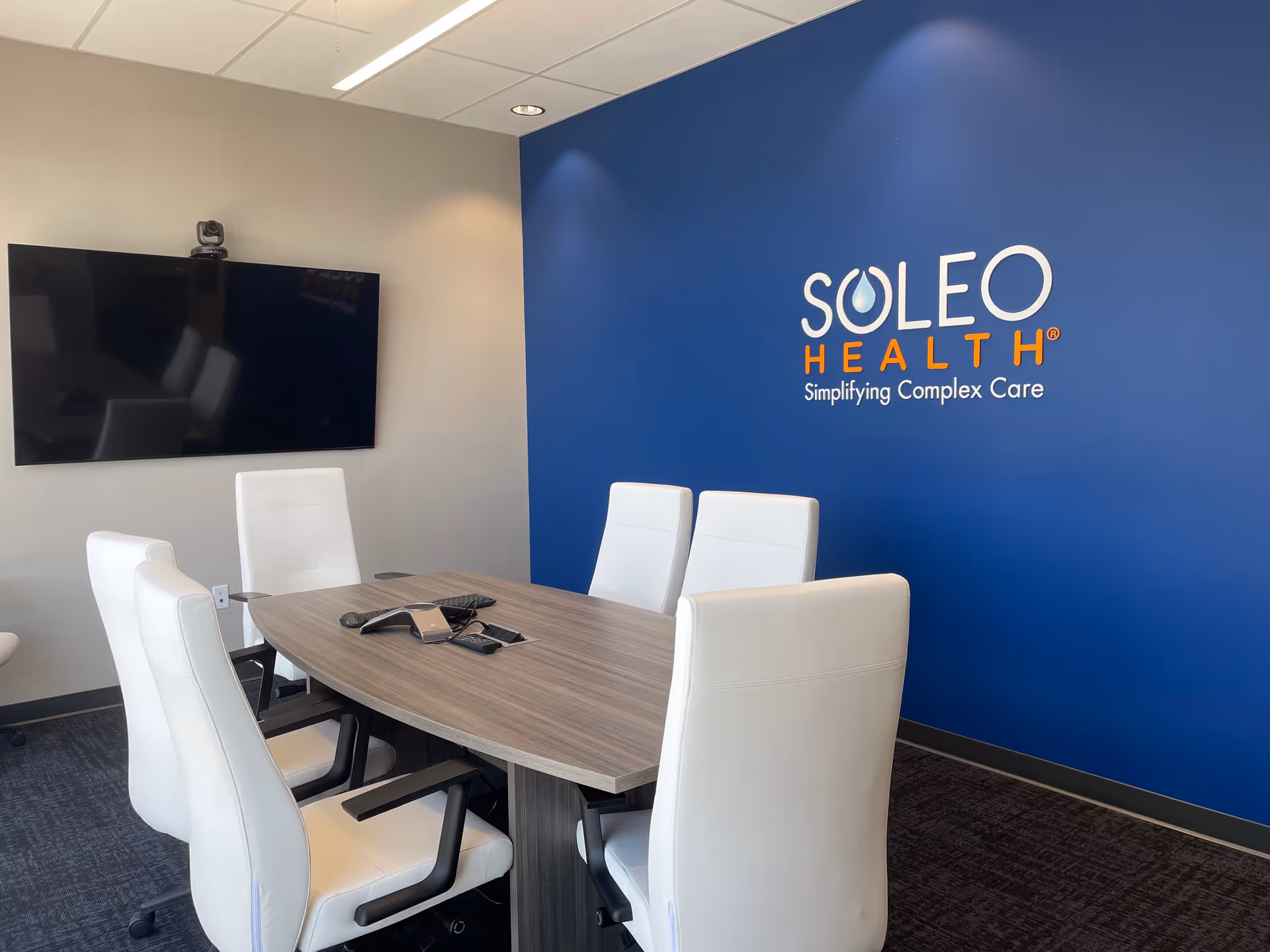 A conference room with a wooden table, white chairs, a wall screen, and Soleo Health logo.