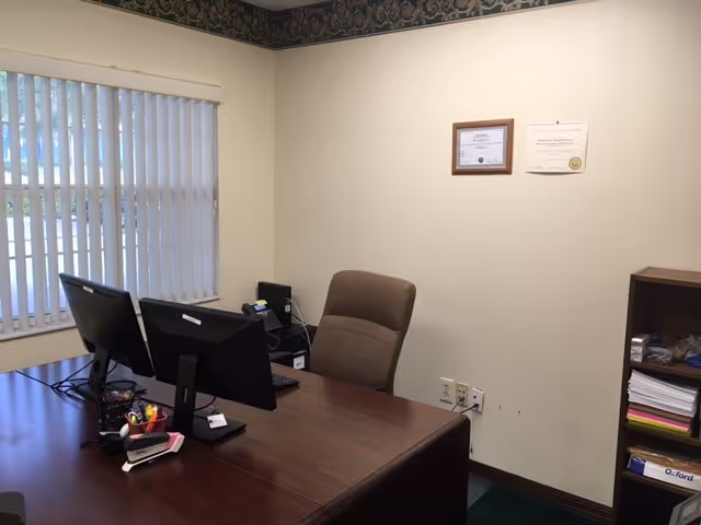 A neat office with a desk, computer, certificate, and shelves.