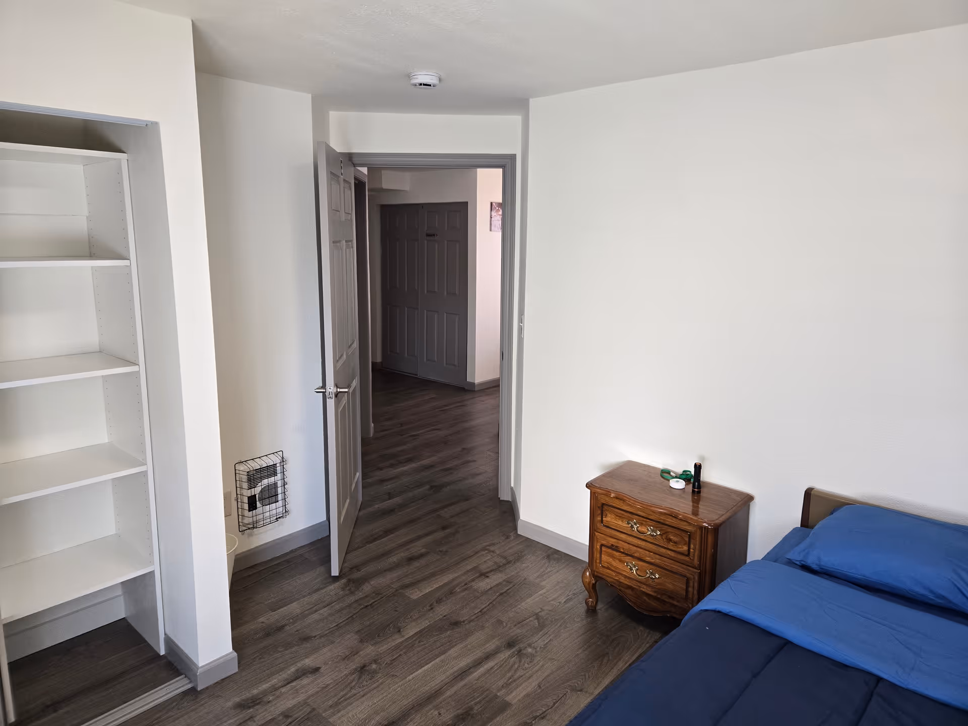 Bedroom in a senior care home featuring a bed, nightstand, bookshelf, and open doorway.