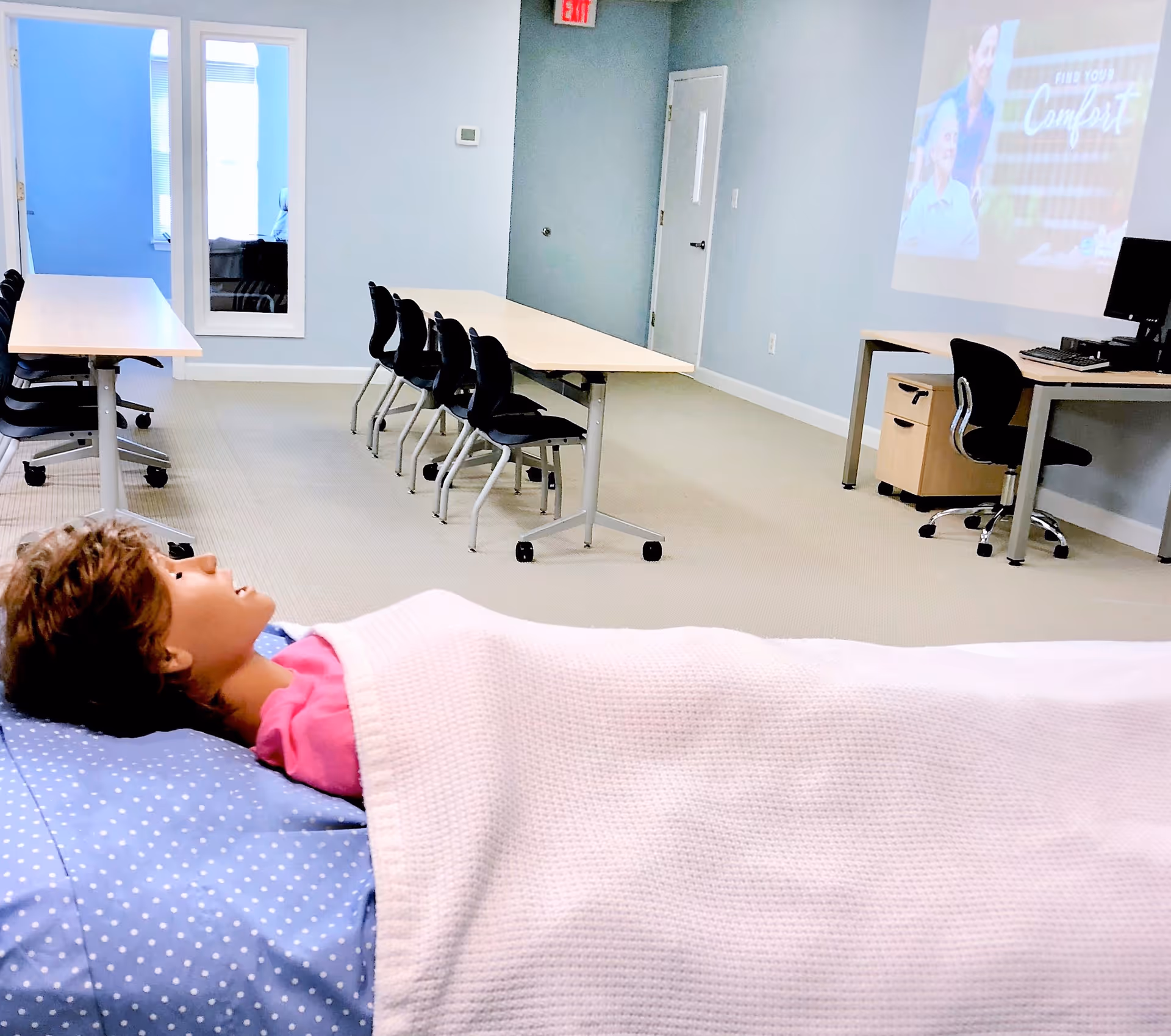 A patient lies in a bed in a bright classroom-like room with tables.