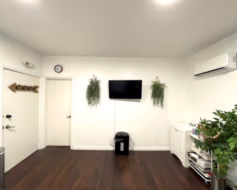 A simple, well-lit lounge area with a wall-mounted TV, plants, and cabinets.
