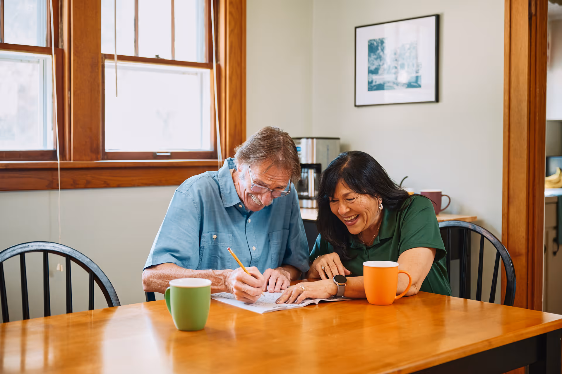 Two seniors sit at a wooden dining table, smiling as they write notes together.