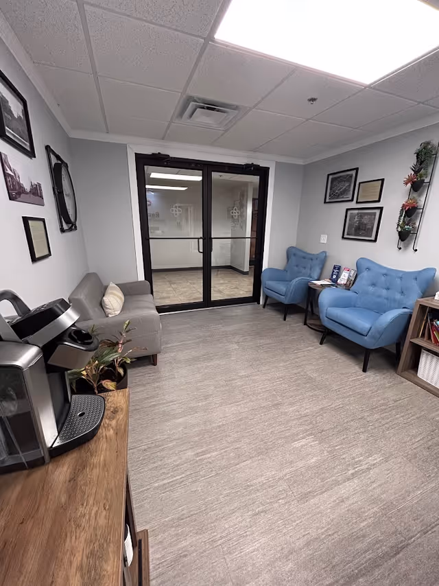 Lobby-style waiting area with gray sofa, blue chairs, coffee station, and glass doors.