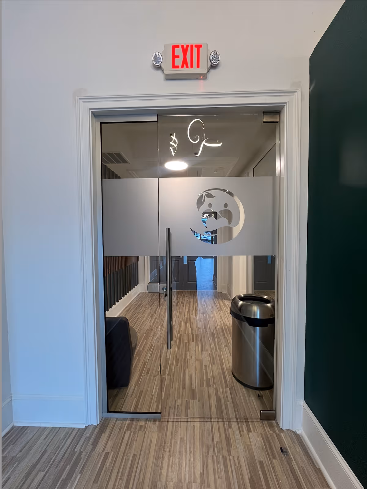 Glass interior door with frosted logo, exit sign above, hallway and trash can inside.