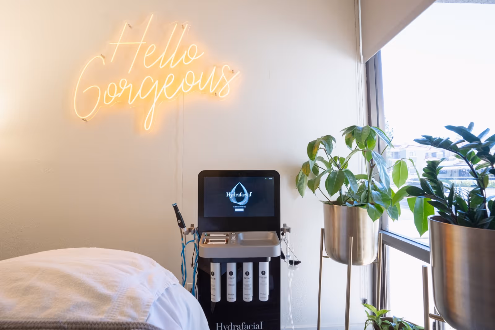 Treatment room with a bed, hydro facial device, neon sign, and tall plants by a window.