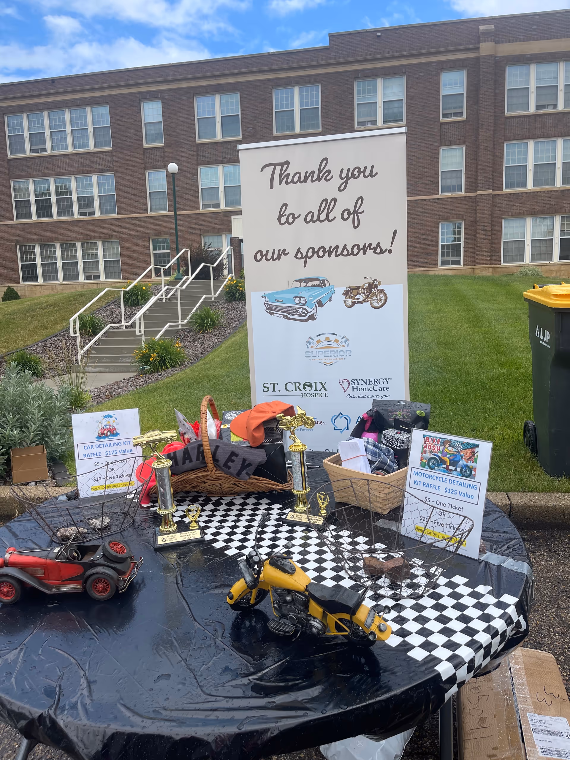 Outdoor sponsor display table with trophies, model cars, and a banner in front of brick facility.