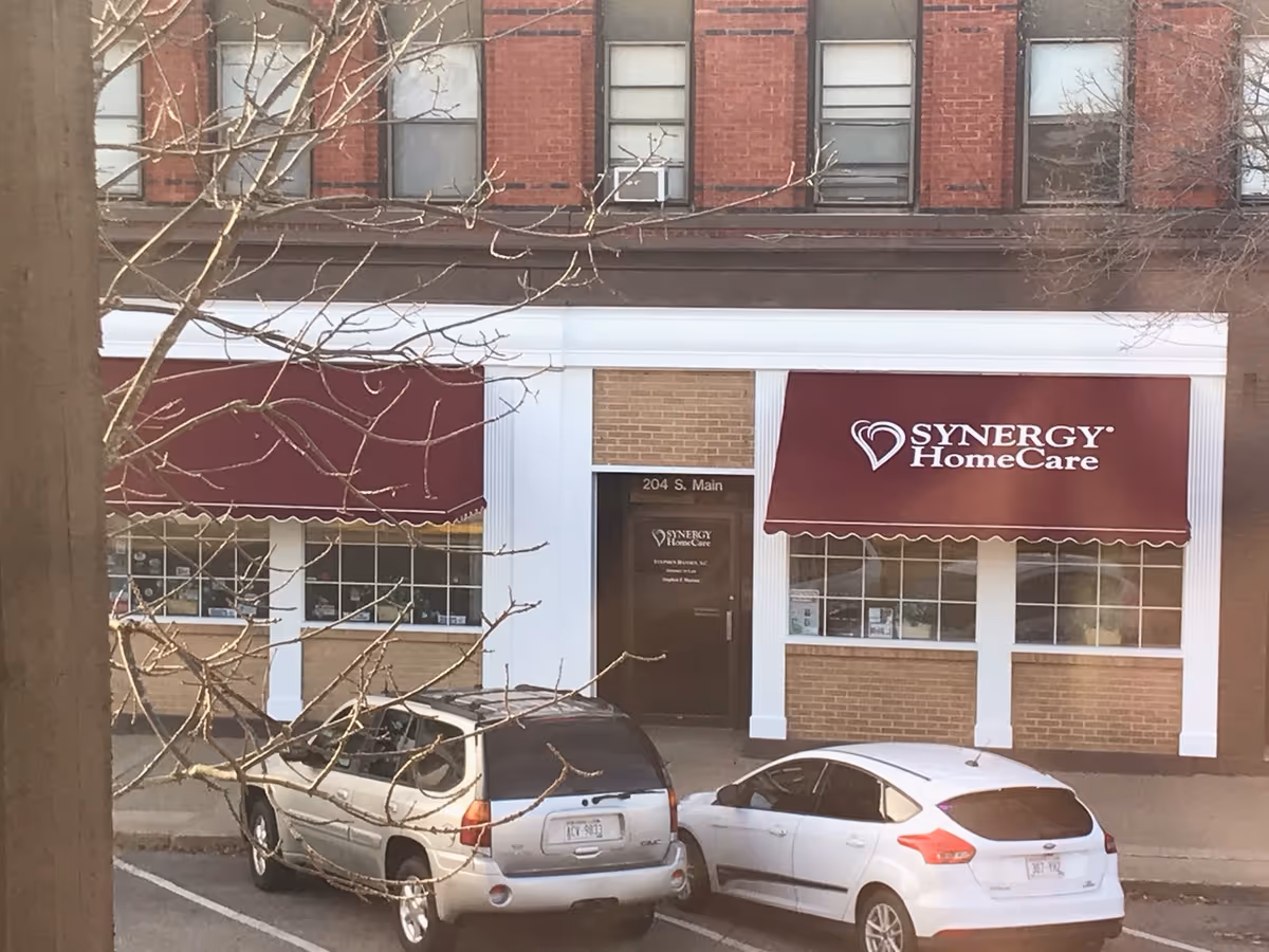 Street view of a SYNERGY HomeCare storefront with maroon awnings and parked cars.