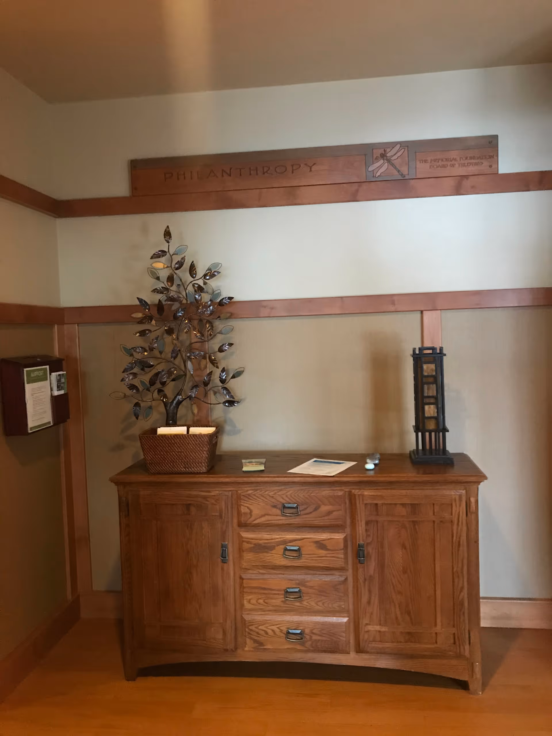 Wooden cabinet with plant and papers beneath a PHILANTHROPHY plaque in a hallway.