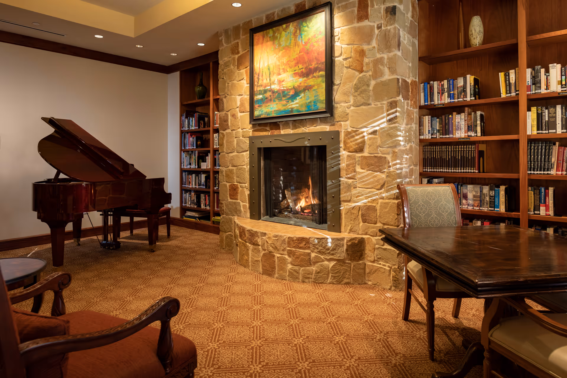Stone fireplace with painting above, surrounded by bookshelves, a grand piano, and a wooden table.