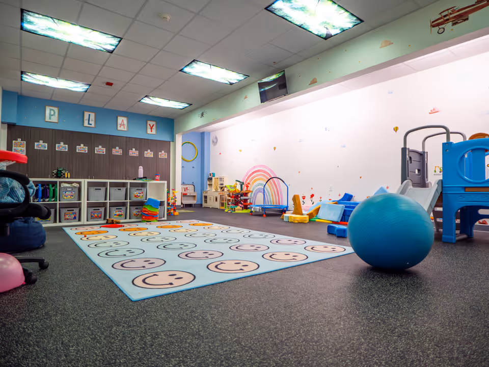 Colorful indoor therapy playroom with mats, toys, a blue exercise ball, and play structures.