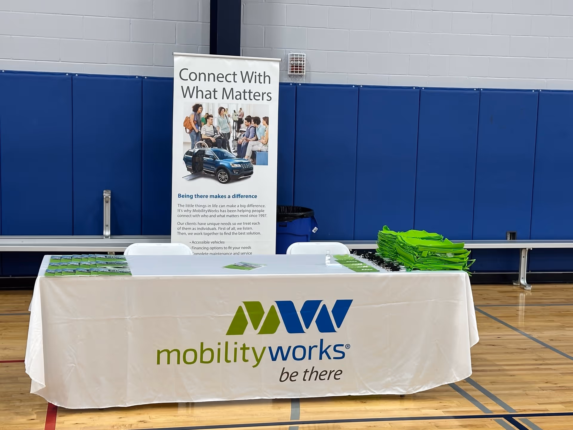 Indoor gym with MobilityWorks booth, banner saying Connect With What Matters, and green folders on table
